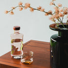 De Paveau Swirl Waterfles Glow Beige 1,25 l van Paveau staat op een houten ondergrond naast een glas water en een vaas met lichtroze bloemen - een ideaal duurzaam cadeau tegen een lichte achtergrond.