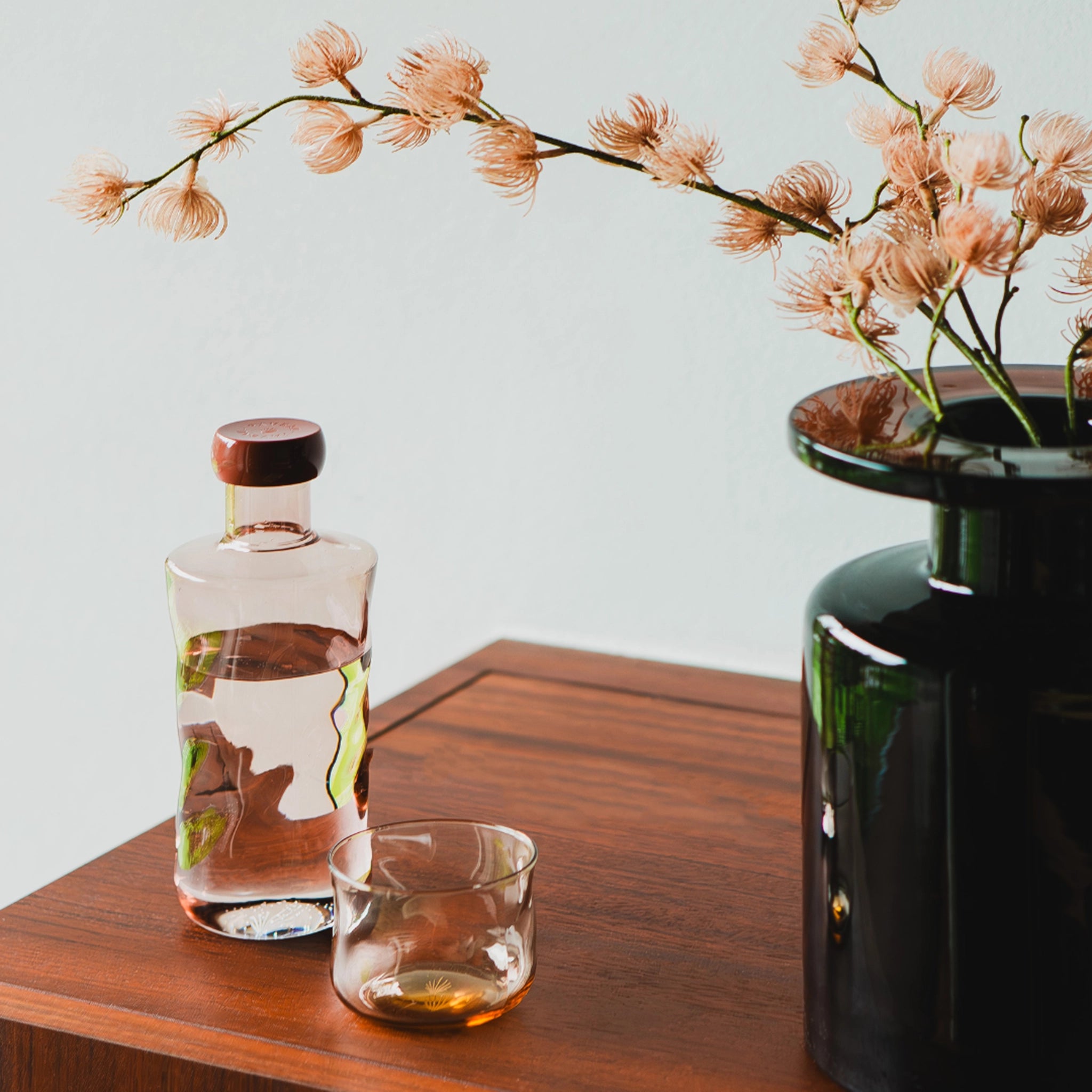 De Paveau Swirl Waterfles Glow Beige 1,25 l van Paveau staat op een houten ondergrond naast een glas water en een vaas met lichtroze bloemen - een ideaal duurzaam cadeau tegen een lichte achtergrond.