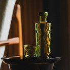 Een groene glazen fles met een bijpassend deksel en een Paveau Swirl Waterglas Moss Groen 24 cl van Paveau rusten op een donkere, glanzende ronde tafel terwijl warm, zacht licht reflecties en schaduwen werpt op de achtergrond.