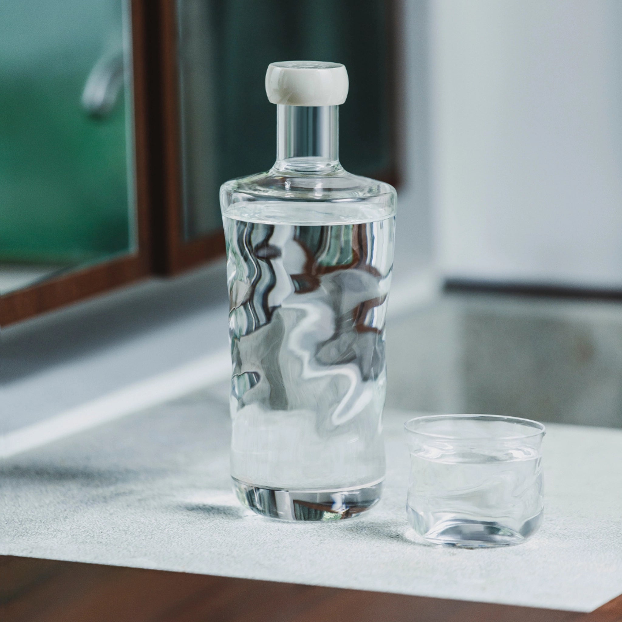 Een glazen karaf met een witte stop staat naast een Paveau Swirl Waterglas Wave Helder 24 cl van Paveau, beide gemaakt van duurzaam glas en geplaatst op een aanrecht in de buurt van een spiegel.