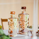 Twee Paveau Swirl Waterflessen Silt Bordeaux 1,25 l van borosilicaatglas met houten doppen staan op een tafel en vangen het licht. De artistieke patronen maken ze tot een stijlvol cadeau tegen een warme, zachte achtergrond.