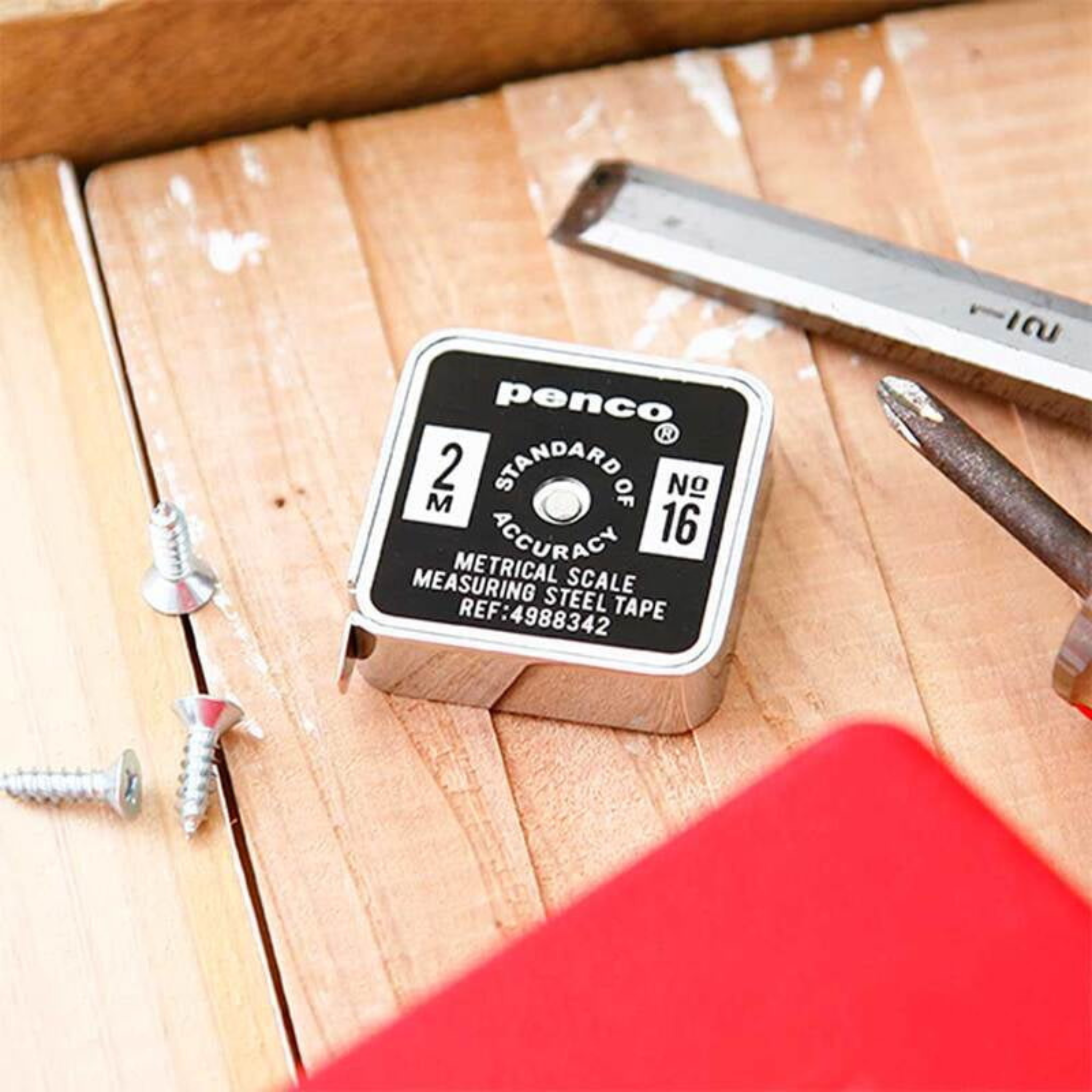 De Penco Compacte Rolmaat Zwart, van het merk Penco, is een handig en compact meetinstrument met een zwart-witte voorkant. Het vierkante ontwerp ligt op hout, naast schroevendraaier, liniaal, schroeven en een rood object.