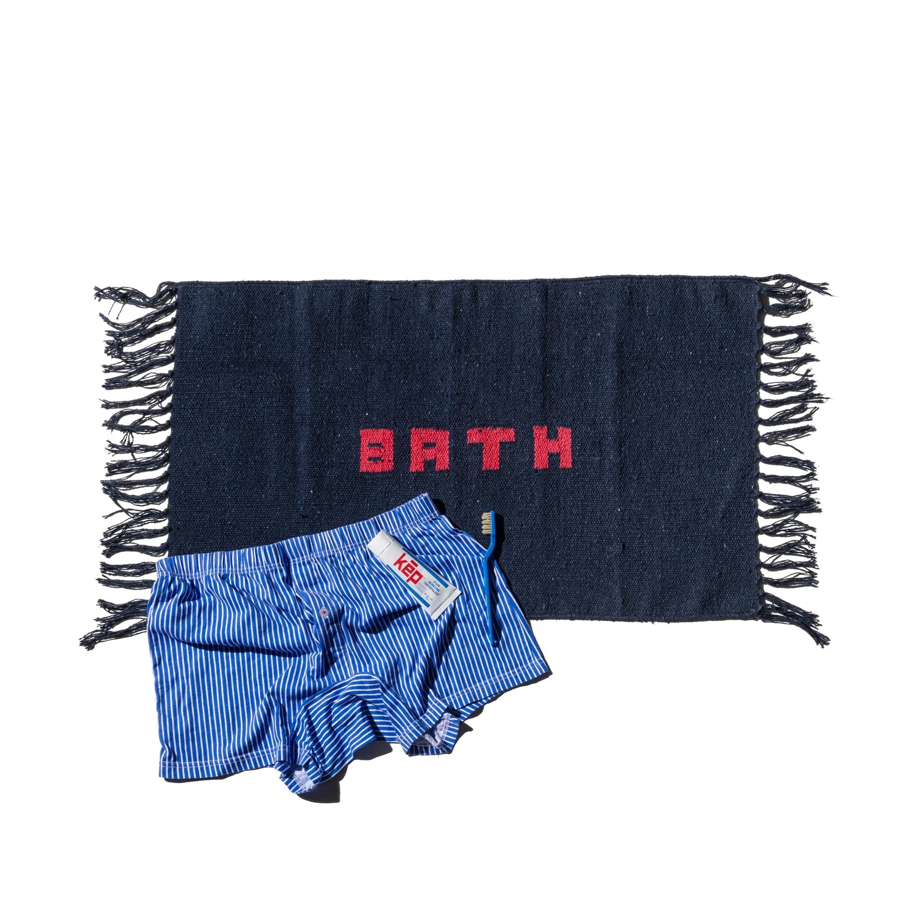 Een Puebco Badmat Gerecycleerd Garen Blauw 75/45 met rode "BATH"-belettering en franje aan de randen wordt gecombineerd met een blauw-wit gestreepte KEY-boxershort, beide getoond op een witte achtergrond.