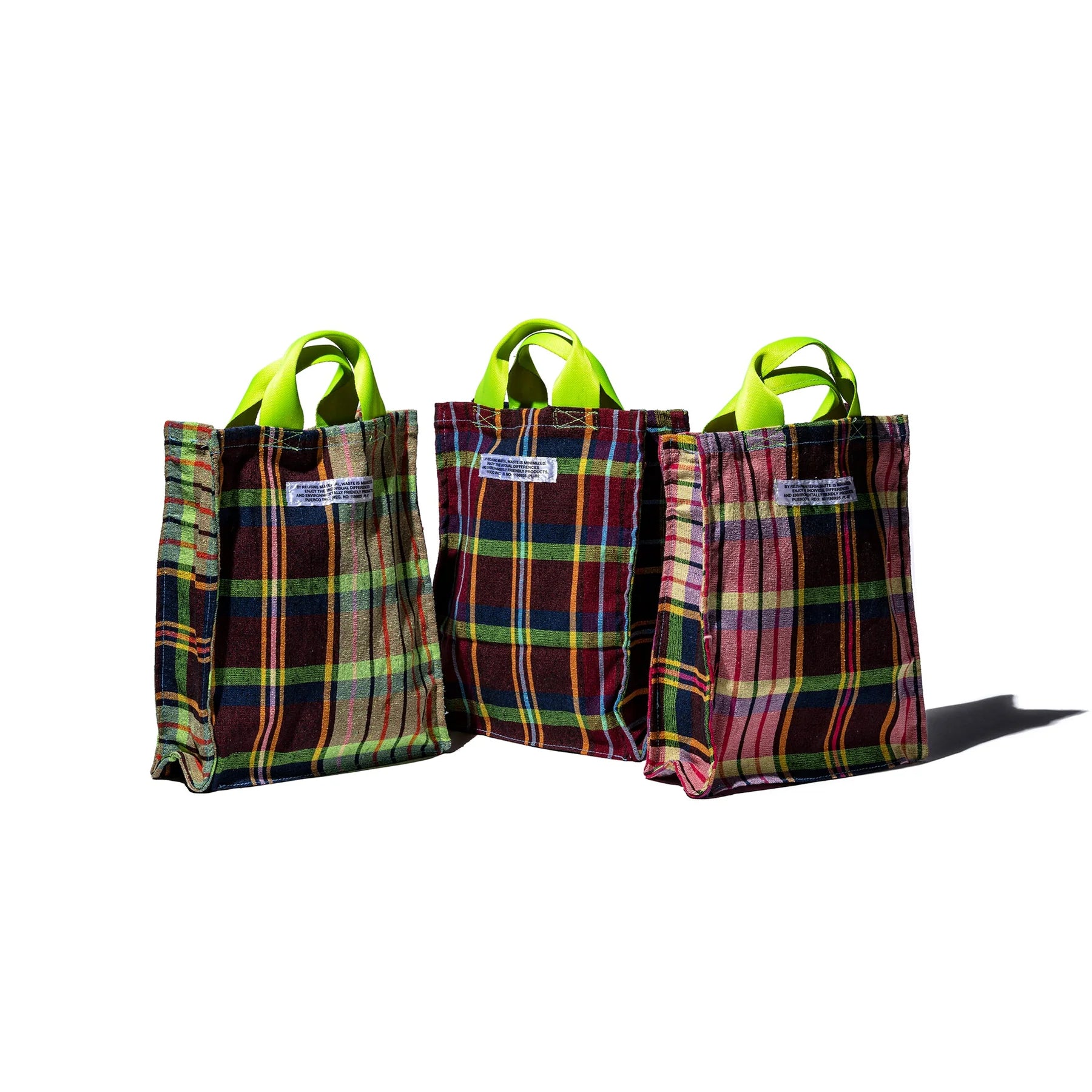 Drie Puebco Check Document Tassen, gemaakt van gerecycleerde stoffen, worden getoond op een witte achtergrond. Elke unieke tas heeft veelkleurige geruite strepen en groene handvatten, met een rechthoekig label op de voorkant.