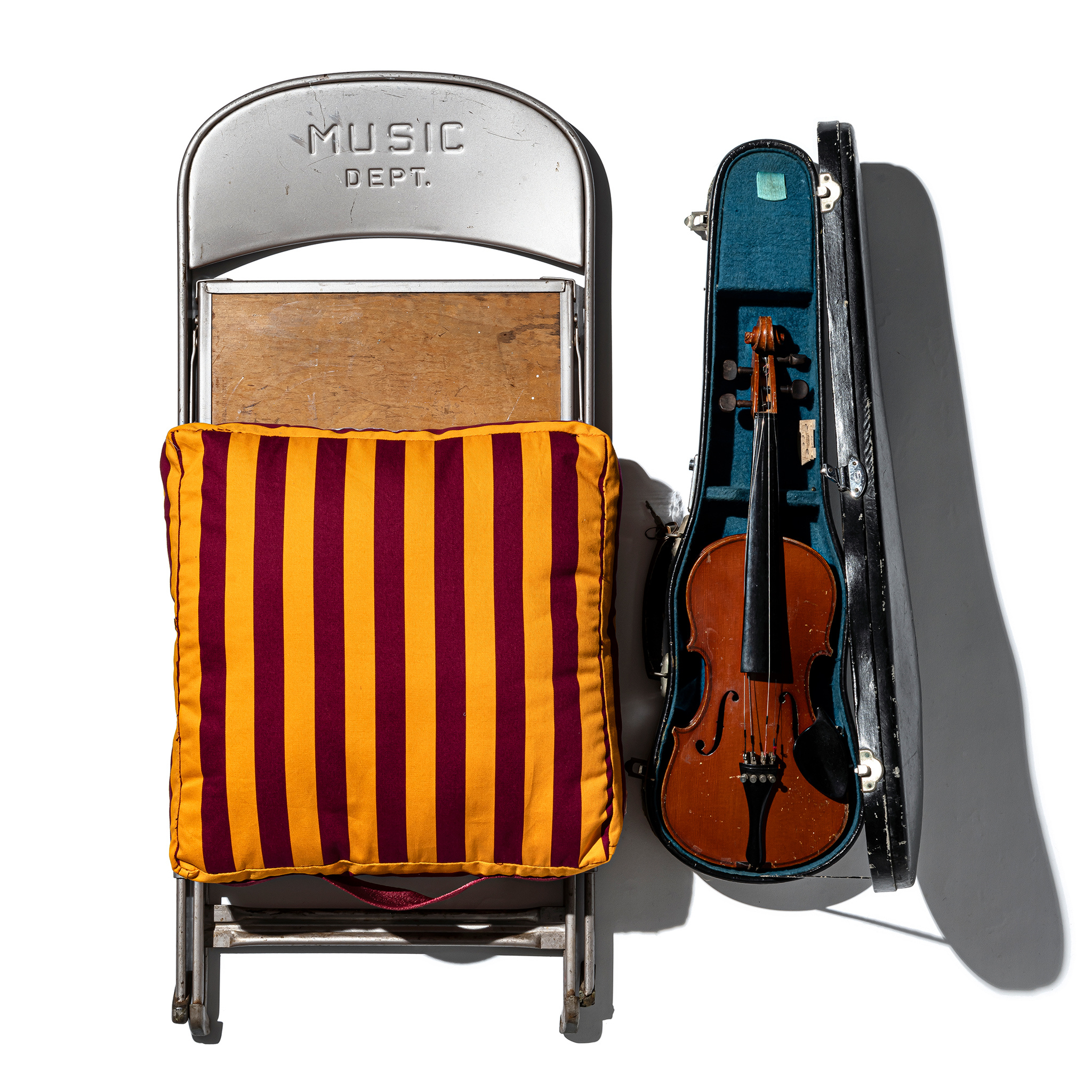 Een metalen klapstoel met het opschrift MUSIC DEPT. en een houten zitting wordt bekleed met een Puebco Draagbaar Kussen Strepen Bordeaux x Mosterd by Puebco voor extra comfort, naast een open vioolkist op een witte achtergrond.