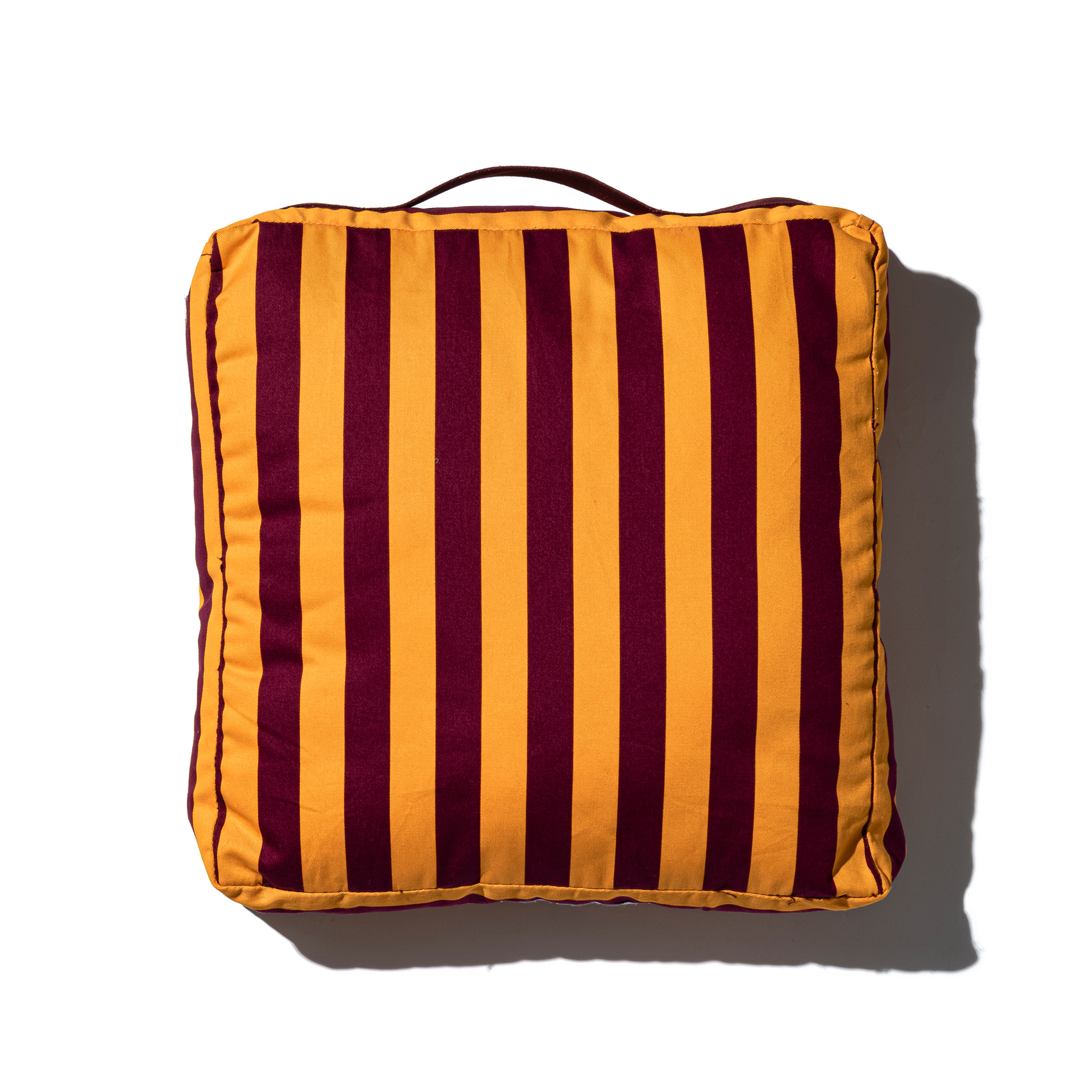 De Puebco Draagbaar Kussen Strepen Bordeaux x Mosterd is een vierkant kussen met een bovenhandgreep, afgebeeld op een witte achtergrond met een schaduw naar rechts. Het biedt extra zitcomfort.