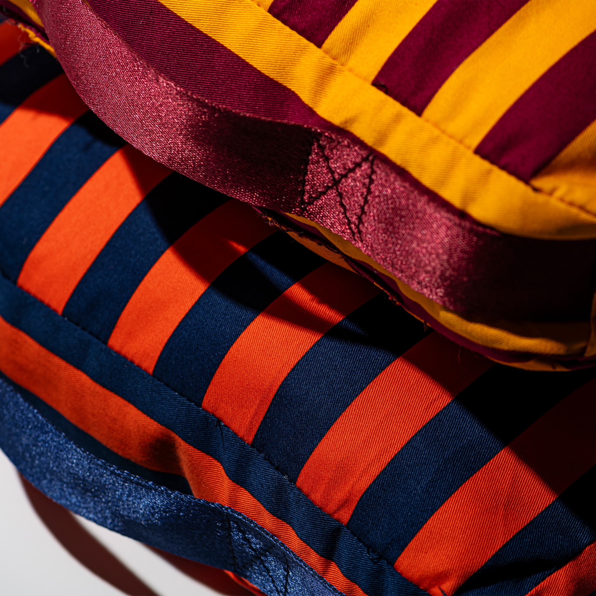 Twee gewatteerde stukken stof met gedurfde strepen: bovenop ligt de Puebco Draagbaar Kussen Strepen Bordeaux x Mosterd voor extra comfort, terwijl het exemplaar eronder oranje en donkerblauwe strepen heeft. Beide hebben gewatteerde, verticale streepontwerpen.