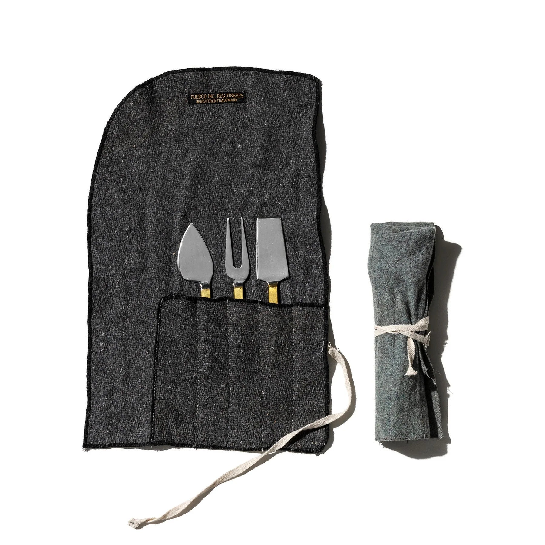 De Puebco Gegraveerde Kaasmesjes 3 st. set van Puebco bestaat uit drie roestvrijstalen kaasmessen en een vork met gouden en zilveren gegraveerde handvatten, in een donkergrijze stoffen etui, open afgebeeld naast dezelfde etui opgerold en dichtgebonden.