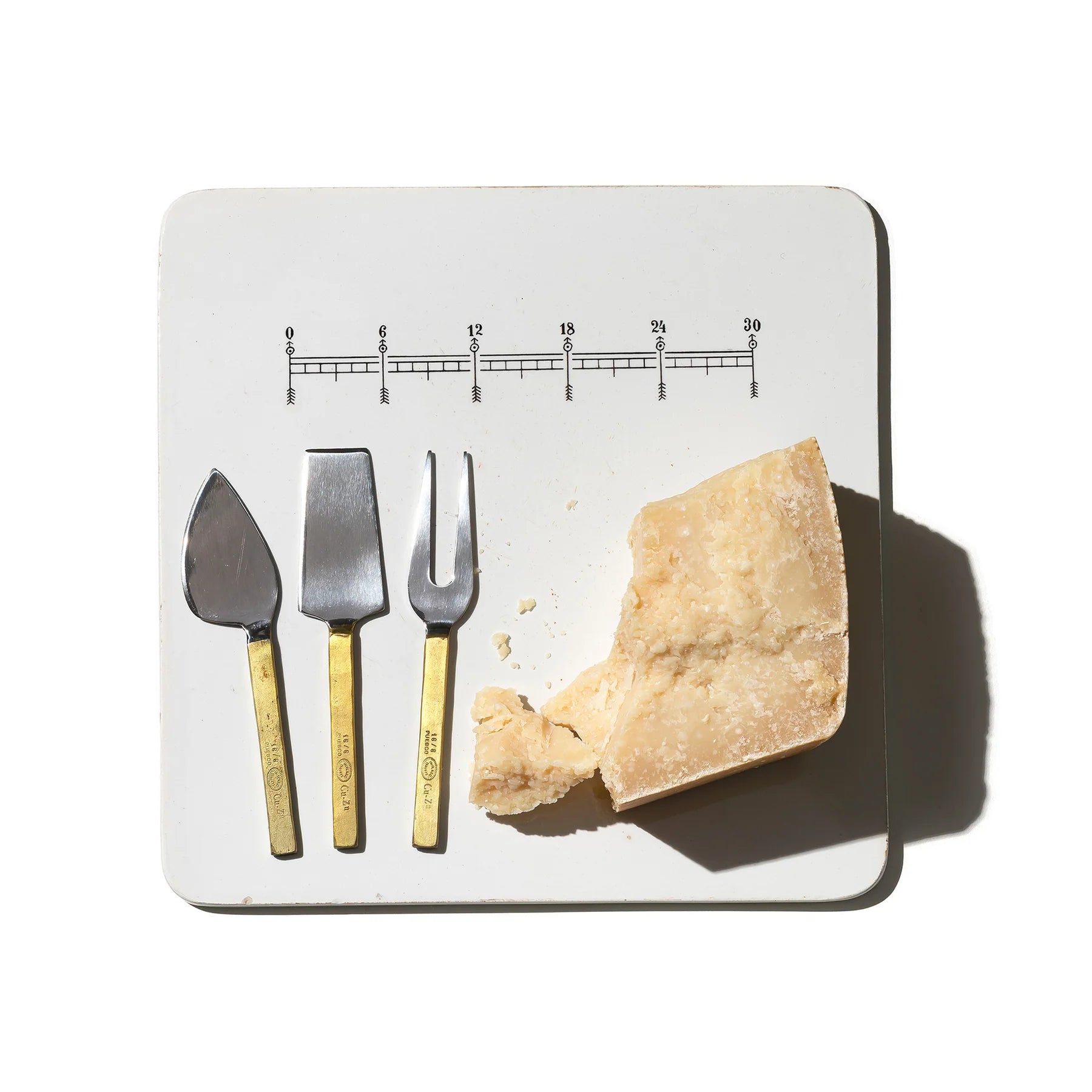 Een blok harde kaas met kruimels ligt op een wit vierkant bord naast Puebco Gegraveerde Kaasmesjes 3 st., elk met gouden handvatten, naast elkaar. Het bord heeft een gegraveerde afbeelding van een liniaal aan de bovenkant.