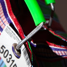 Close-up van een metalen karabijnhaak gegraveerd met PUEBCO INC., bevestigd aan de Puebco Gerecycleerde Tas Strepen Oranje Roze D26. Aan deze kleurrijke Puebco-tas hangt een wit label met zwarte cijfers en een streepjescode.