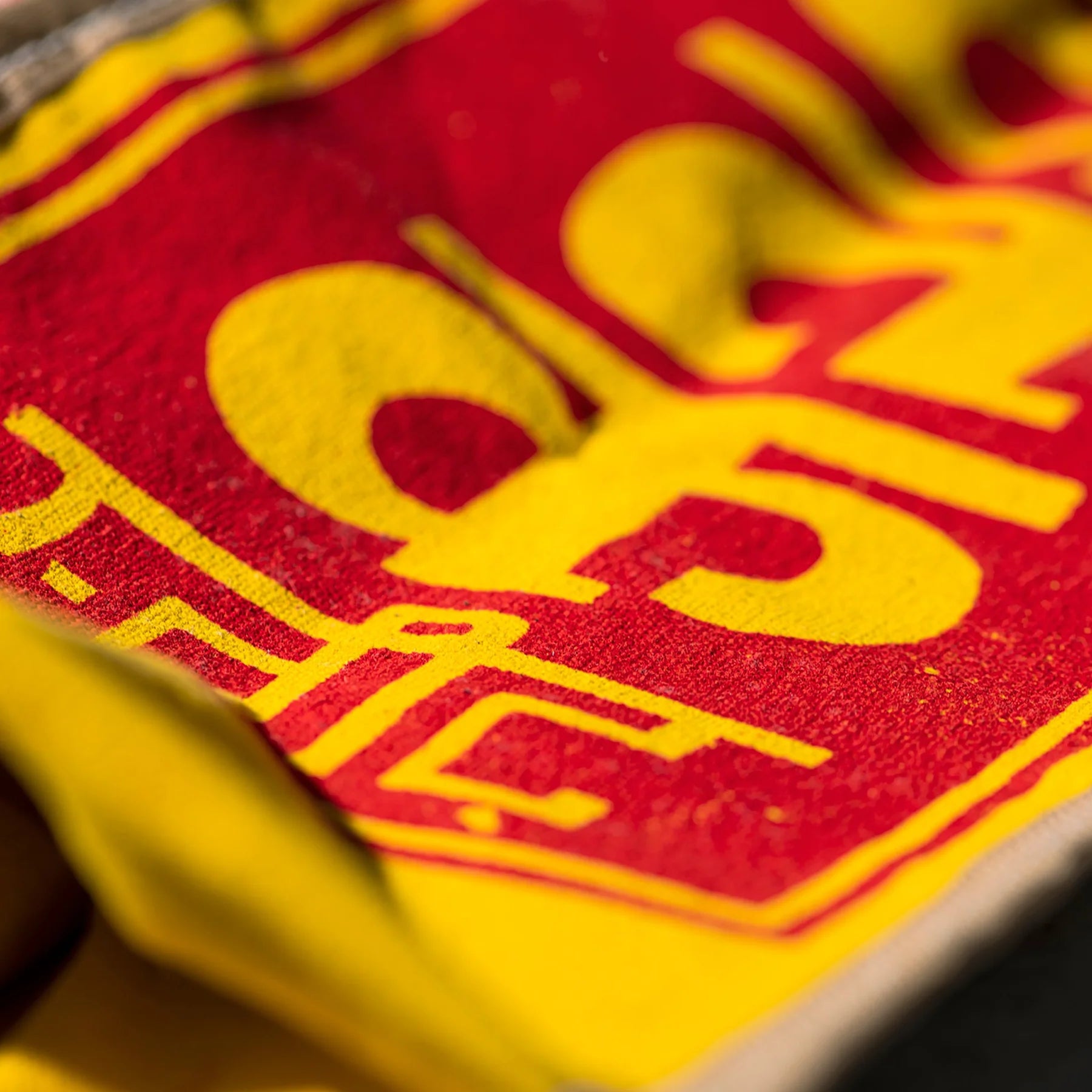 Close-up van een Puebco India Souvenir Tas stof met opvallende rode en gele print met grote, gedeeltelijk zichtbare letters en cijfers, waardoor een abstract, kleurrijk patroon op een gestructureerd oppervlak ontstaat.