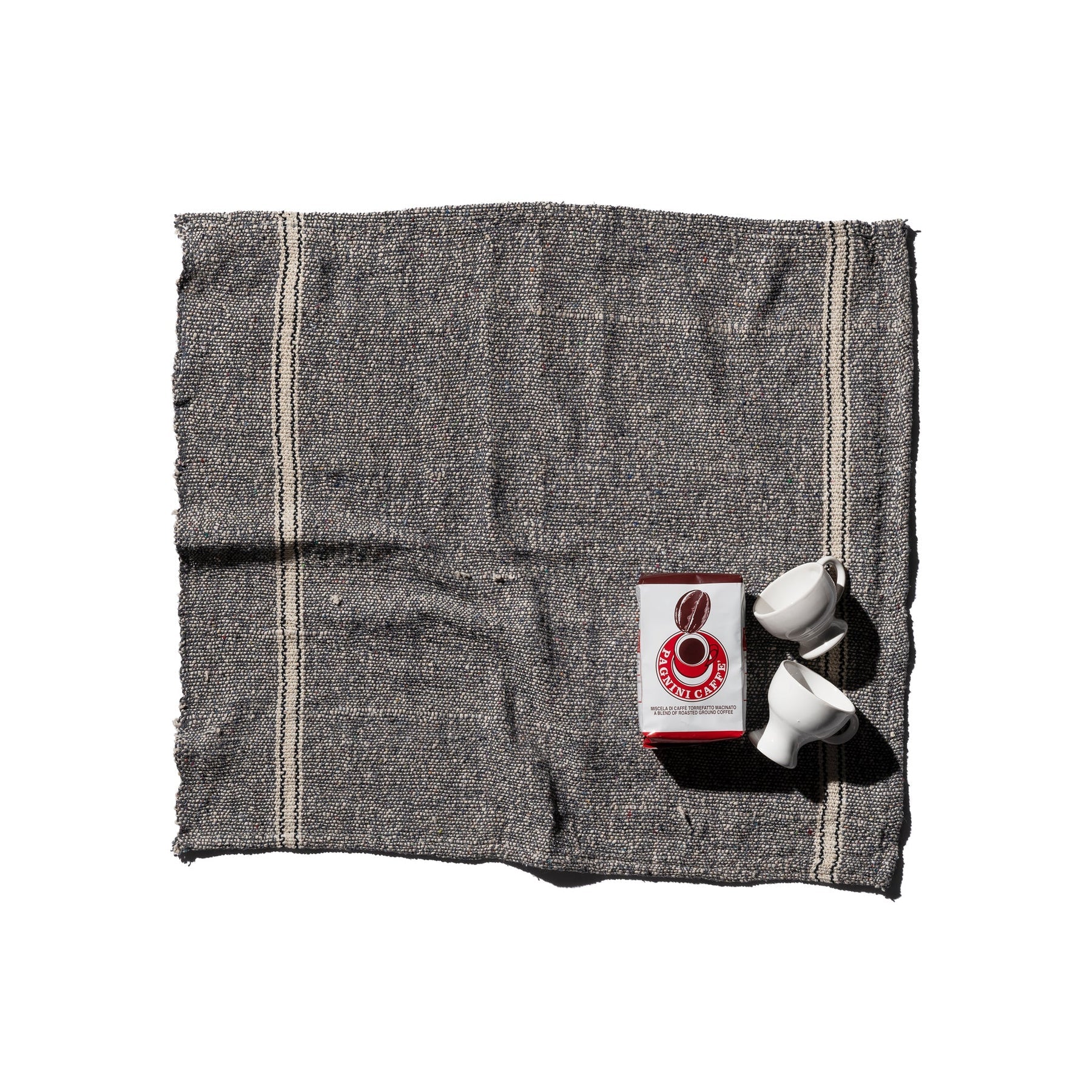De Puebco India Theedoek Grijs 60/60 van Puebco, vervaardigd uit gerecycleerde stoffen, vormt het decor voor twee witte koffiekopjes en een rood-witte Counter Culture Coffee verpakking tegen een witte achtergrond.