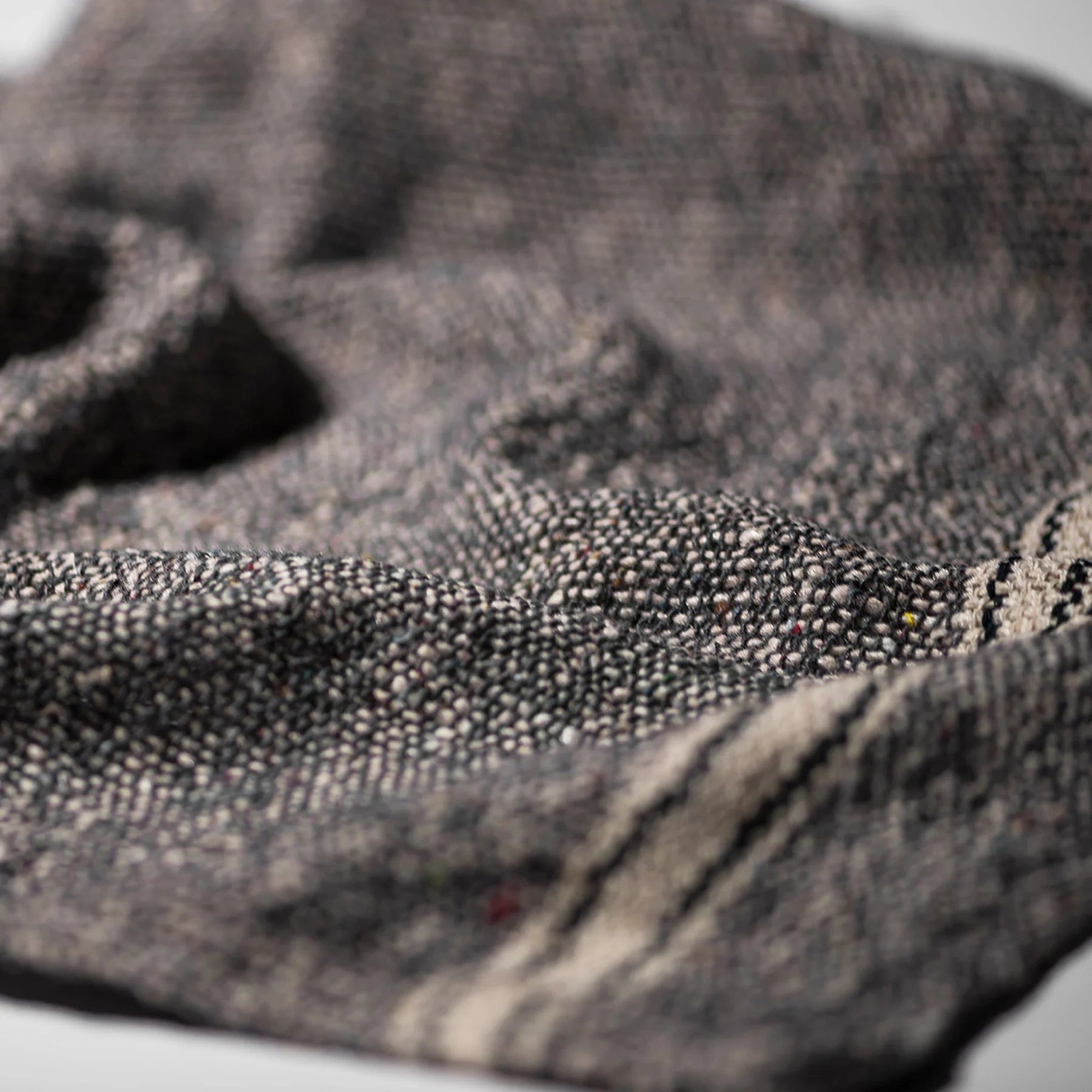 Een close-up van de Puebco India Theedoek Grijs 60/60, een grijze geweven stof van Puebco, met een crèmekleurige streep in de rechterbenedenhoek. De zachte stof is gemaakt van gerecycleerde stoffen en heeft een licht gerimpelde textuur.