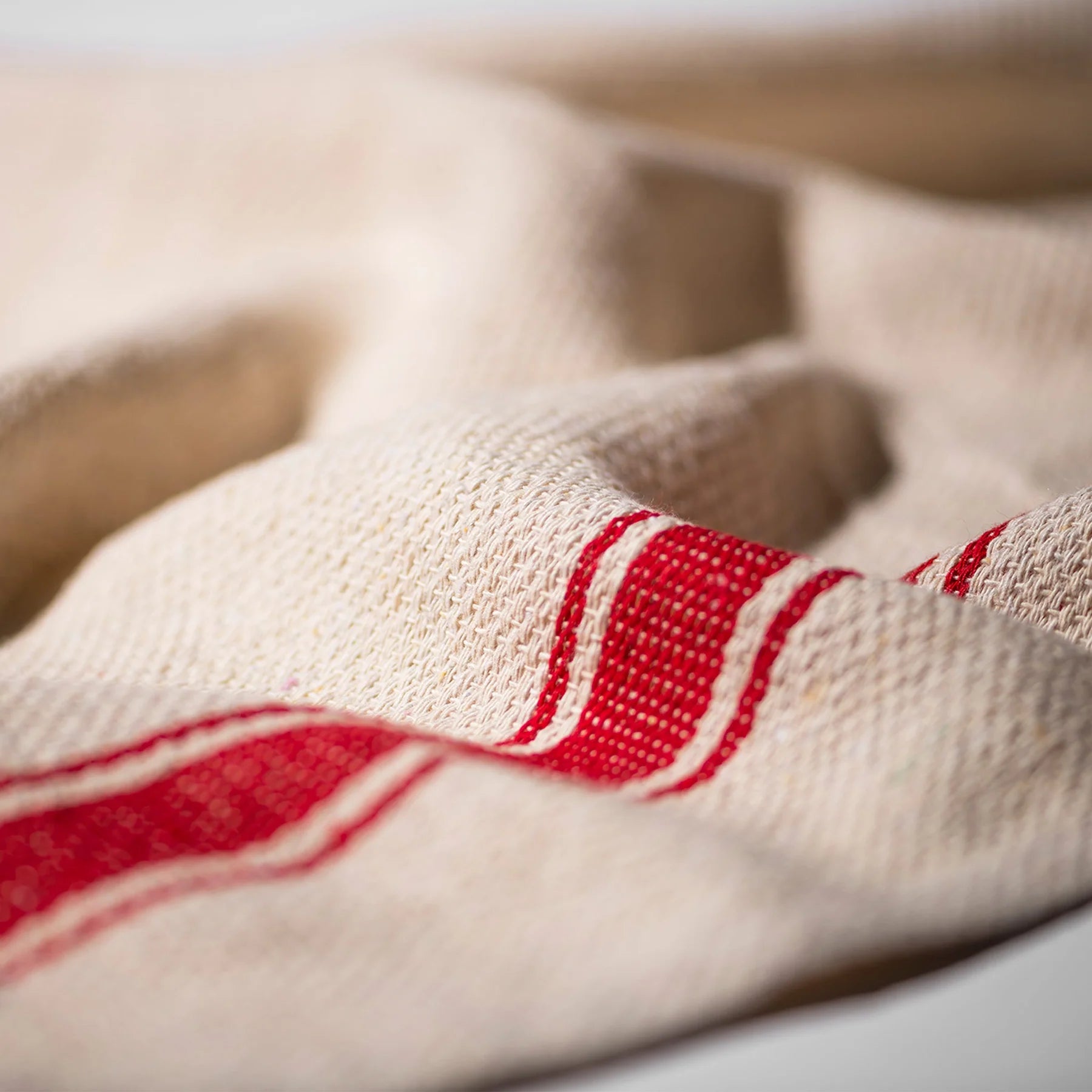 Een close-up van de Puebco India Theedoek Rood 60/60 van Puebco toont de beige stof met textuur en twee rode strepen, fijne weefdetails, zachte plooien en milieuvriendelijke constructie van gerecyclede materialen.