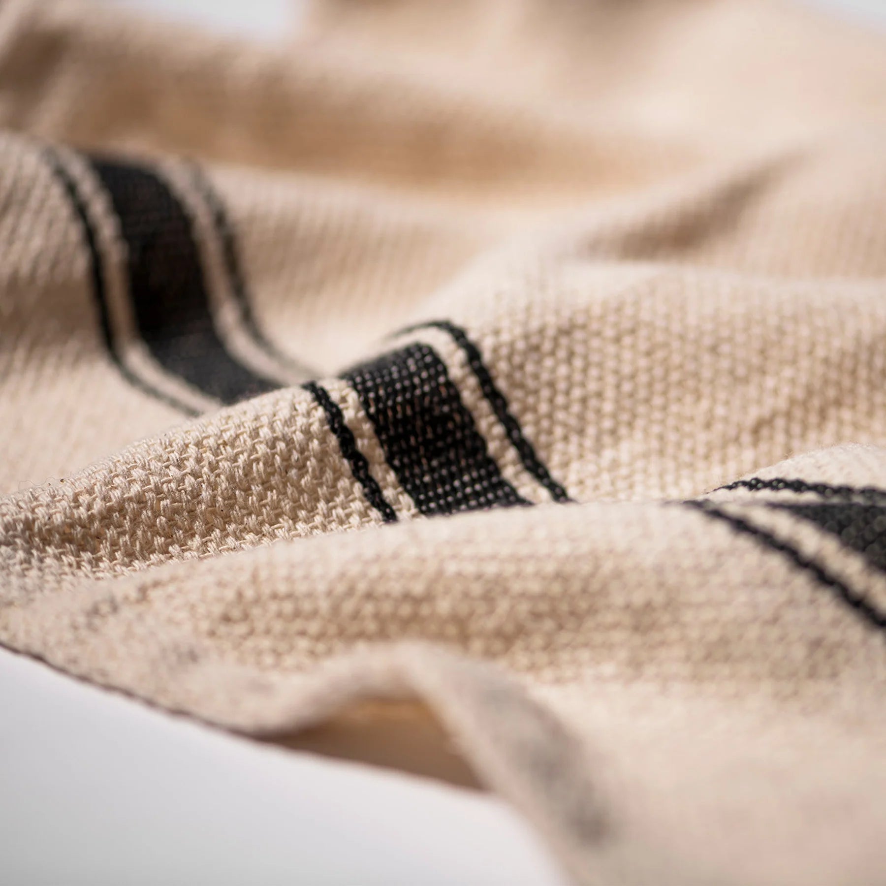 Een close-up van de Puebco India Theedoek Zwart 60/60 toont de beige stof met textuur en parallelle zwarte strepen, die het geweven patroon en de zachte plooien van gerecycled materiaal benadrukken. Merk: Puebco.