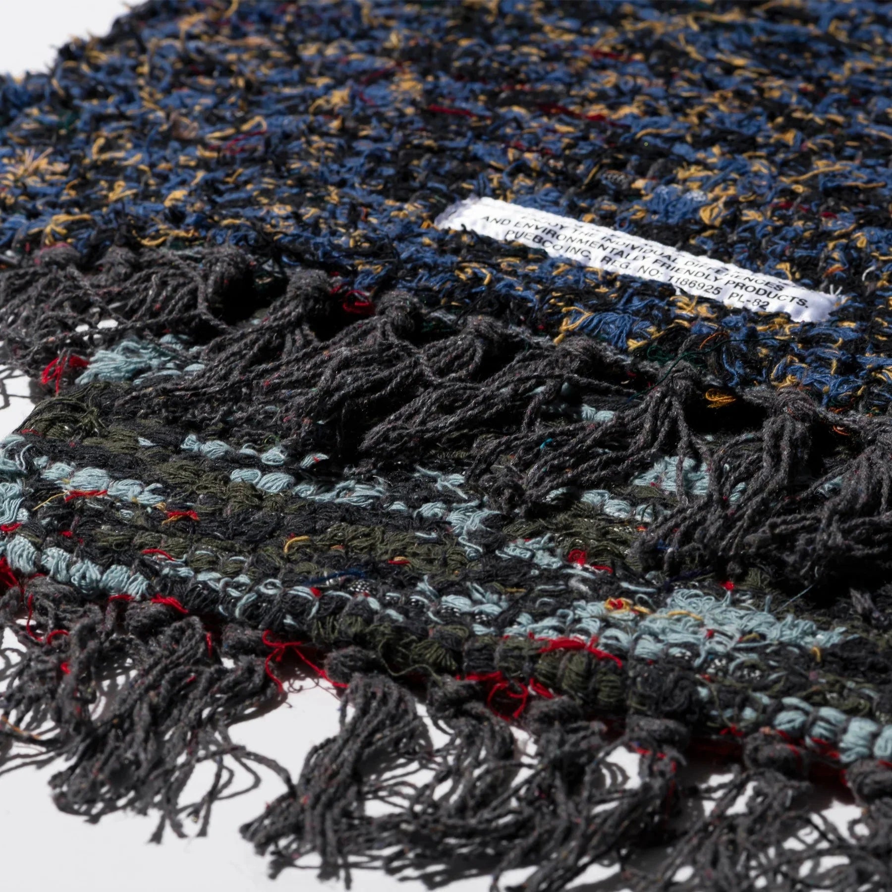 Een close-up van de Puebco Mat Gerecycleerd Textiel 63/41 van Puebco, met de veelkleurige geweven draden-blauw, zwart, rood, geel-omzoomde randen en een klein wit label met gedeeltelijk zichtbare tekst.