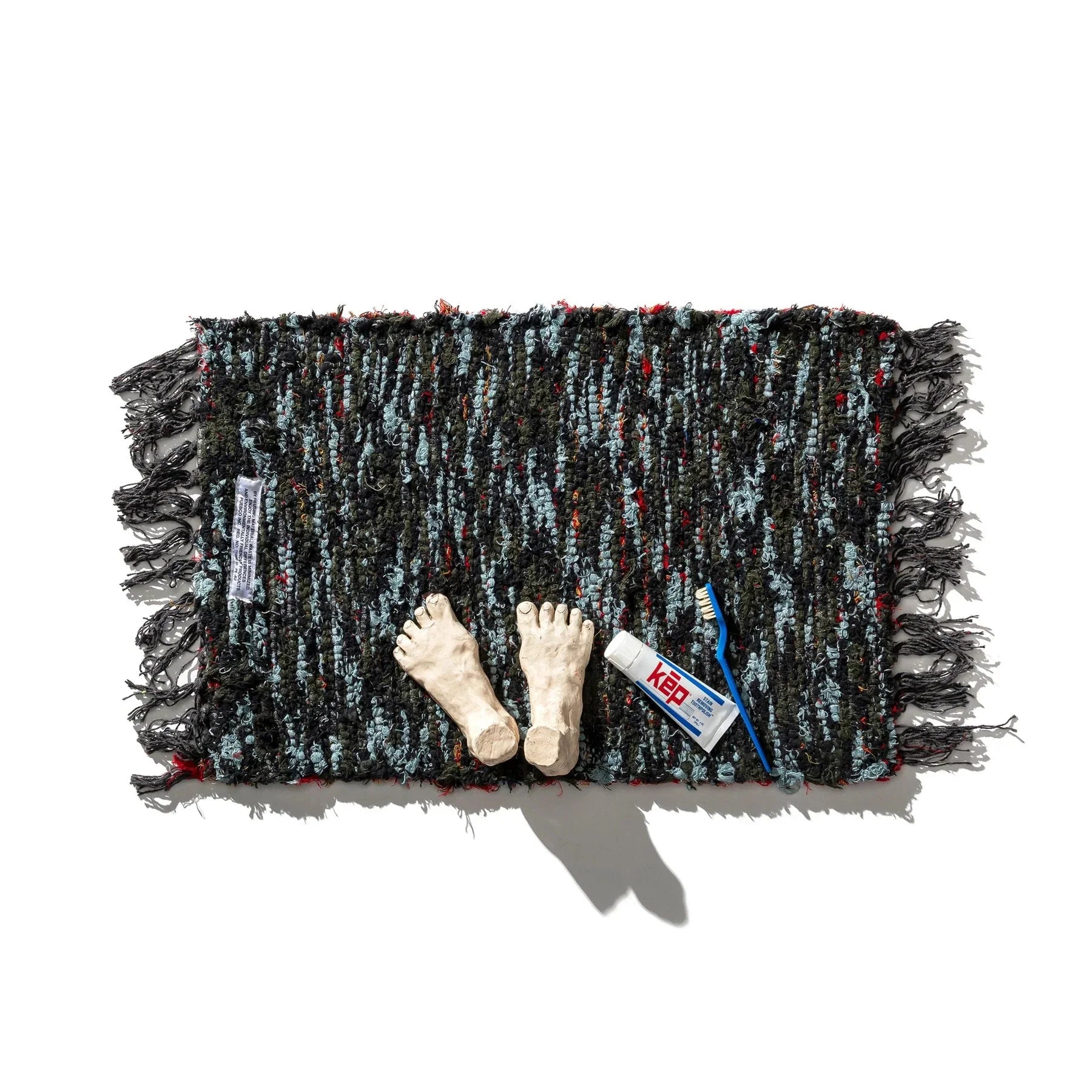 De Puebco Mat Gerecycleerd Textiel 63/41, een mat met donkere structuur en franjes van Puebco, is afgebeeld op een witte achtergrond met daarop twee modelvoeten, een tube Keb tandpasta en een blauwe tandenborstel.