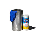 Een paar blauwe wegwerphandschoenen ligt op een grote Puebco Prullenbak Gerecycleerd Staal H28 naast een container met ontsmettingsdoekjes van Clorox, alles op een witte achtergrond.
