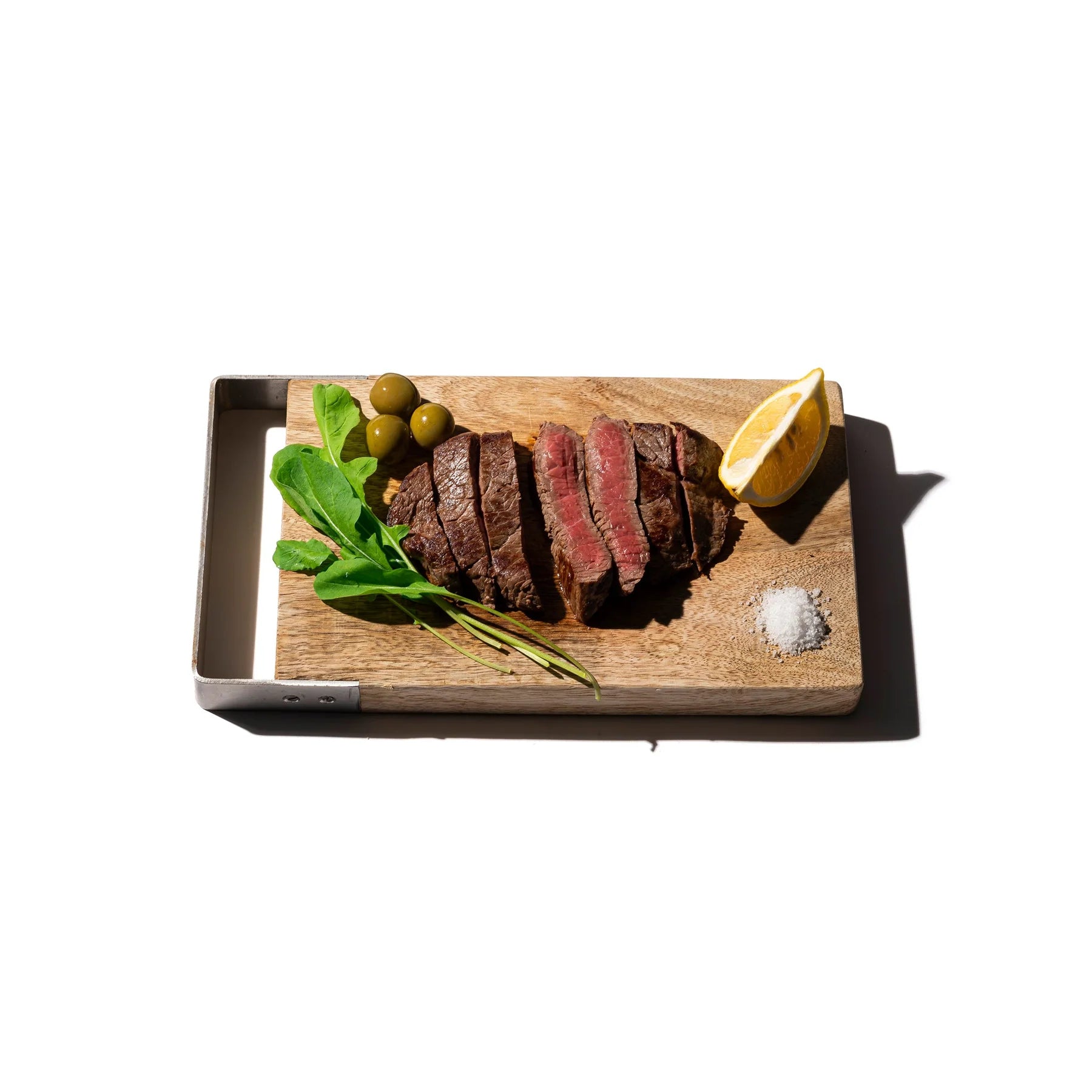 Gesneden biefstuk wordt geserveerd op de Puebco Snijplank Mango Hout 17/27, vergezeld van groene bladgroenten, groene olijven, een citroenpartje en een klein hoopje zout, alles tegen een witte achtergrond.