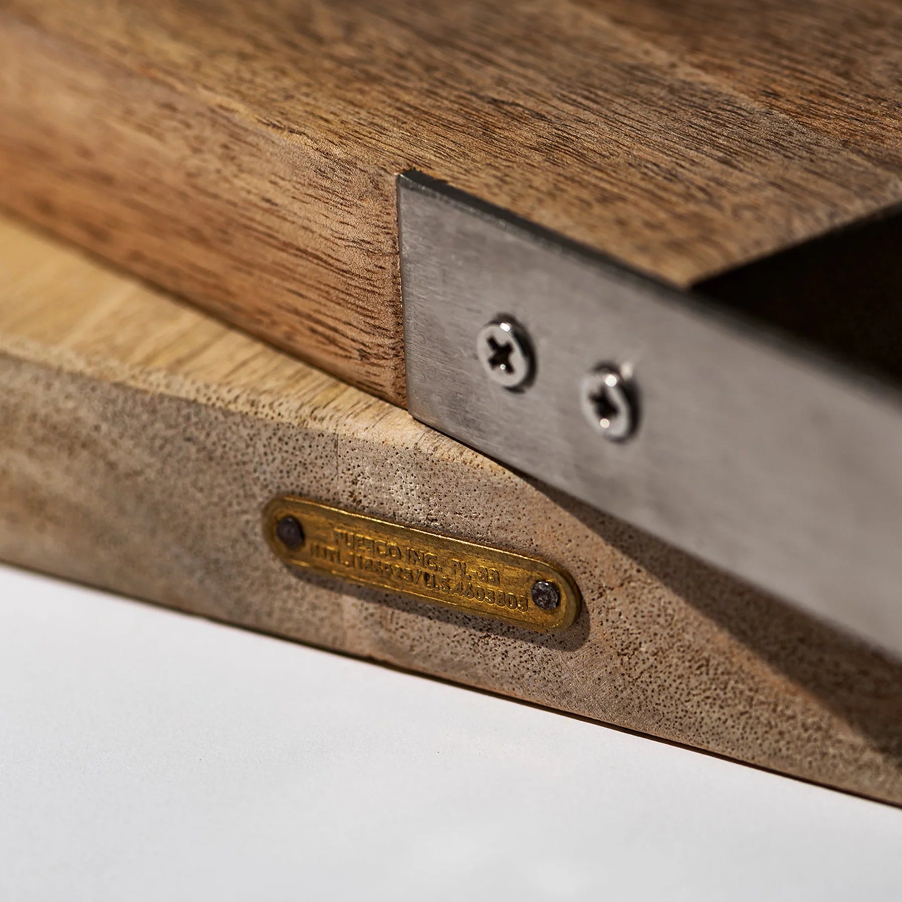 Close-up van de Puebco Snijplank Mango Hout 20/32 van Puebco, met een stalen handvat en metalen plaat bevestigd met twee schroeven. Een klein gegraveerd messing naamplaatje benadrukt de unieke natuurlijke nerf van het mangohout.