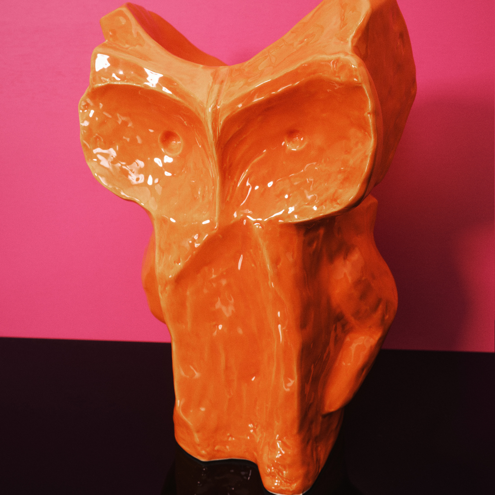 De Serax Marni After Midnight Uil Vaas Oranje S, een glanzend abstract oranje keramische uil sculptuur, wordt getoond tegen een roze en donkerpaarse achtergrond.