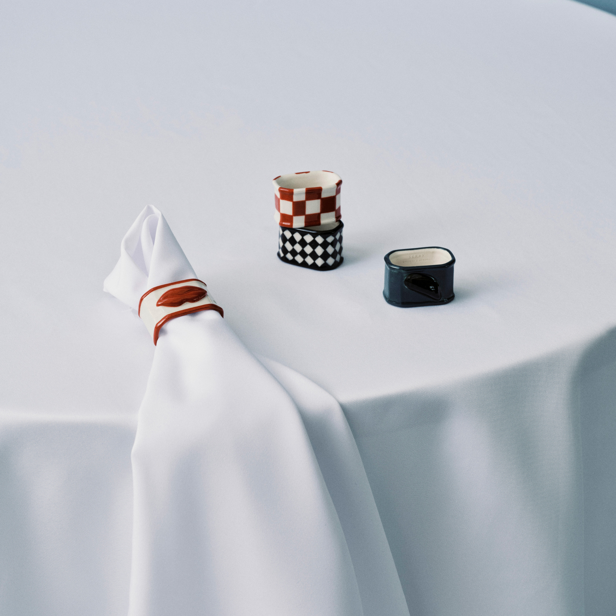 Een wit tafelkleed bedekt een ronde tafel. Bovenop ligt een gevouwen wit servet met een Serax Wouters & Hendrix servetring, een klein kopje met rode en zwarte motieven en een blauw vierkant bakje met een zwart deksel.
