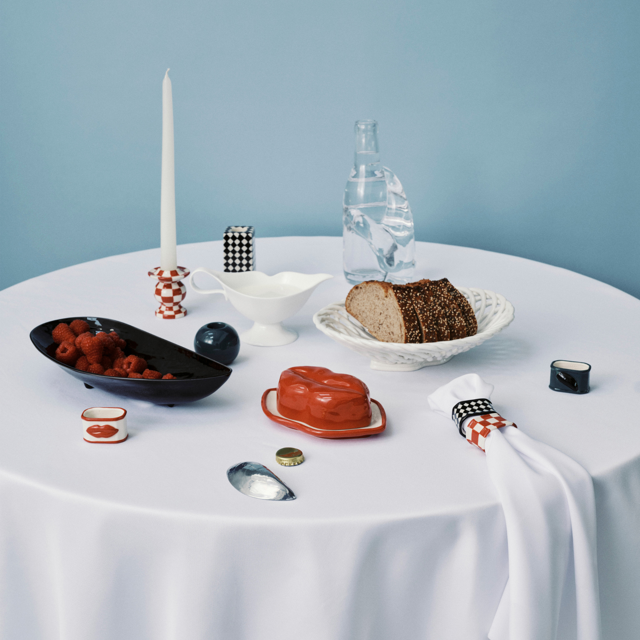 Een ronde witte tafel met brood, frambozen, een rood botervlootje, een kaars, een waterfles en een elegant decor - benadrukt door de Serax Wouters & Hendrix Servetring 4 st. tegen een lichtblauwe achtergrond.