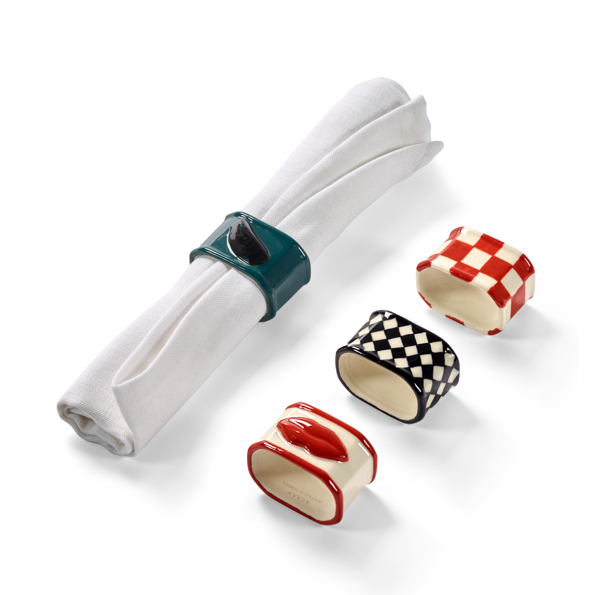 Een wit opgerold servet wordt vastgehouden door een groene Serax Wouters & Hendrix servetring in de vorm van een kat. Daarnaast liggen drie servetringen van keramiek van Serax: een rood-wit geruite, een zwart-wit geruite en een in de vorm van rode lippen.