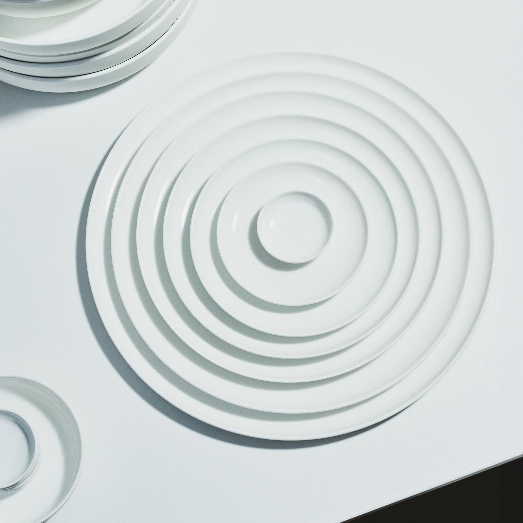 Een stapel witte Serax Base Laag Bord D20 borden vormt geneste cirkels op een wit oppervlak. Hun ronde vorm en verschillende afmetingen weerspiegelen de elegantie van Chinees porselein. Extra Serax borden zijn gedeeltelijk zichtbaar aan de randen.