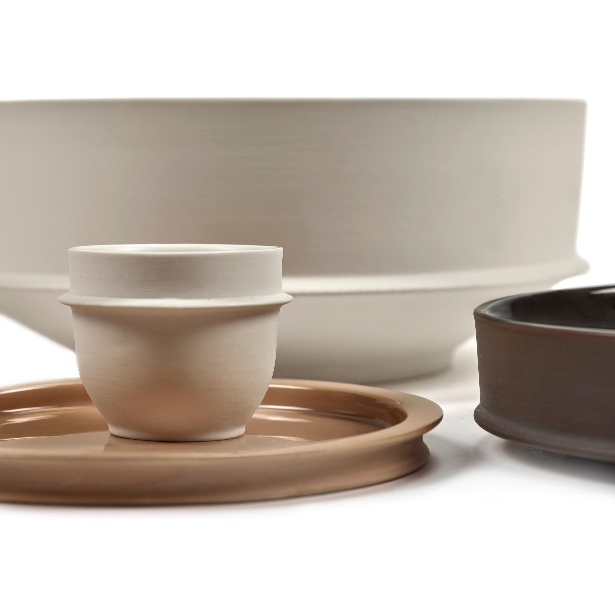 Een set minimalistisch servies van keramiek, met het Serax Dune Laag Bord Clay D17 bord, een witte kom, een kleine beige beker en een donkere kom, staan dicht op elkaar op een witte achtergrond.
