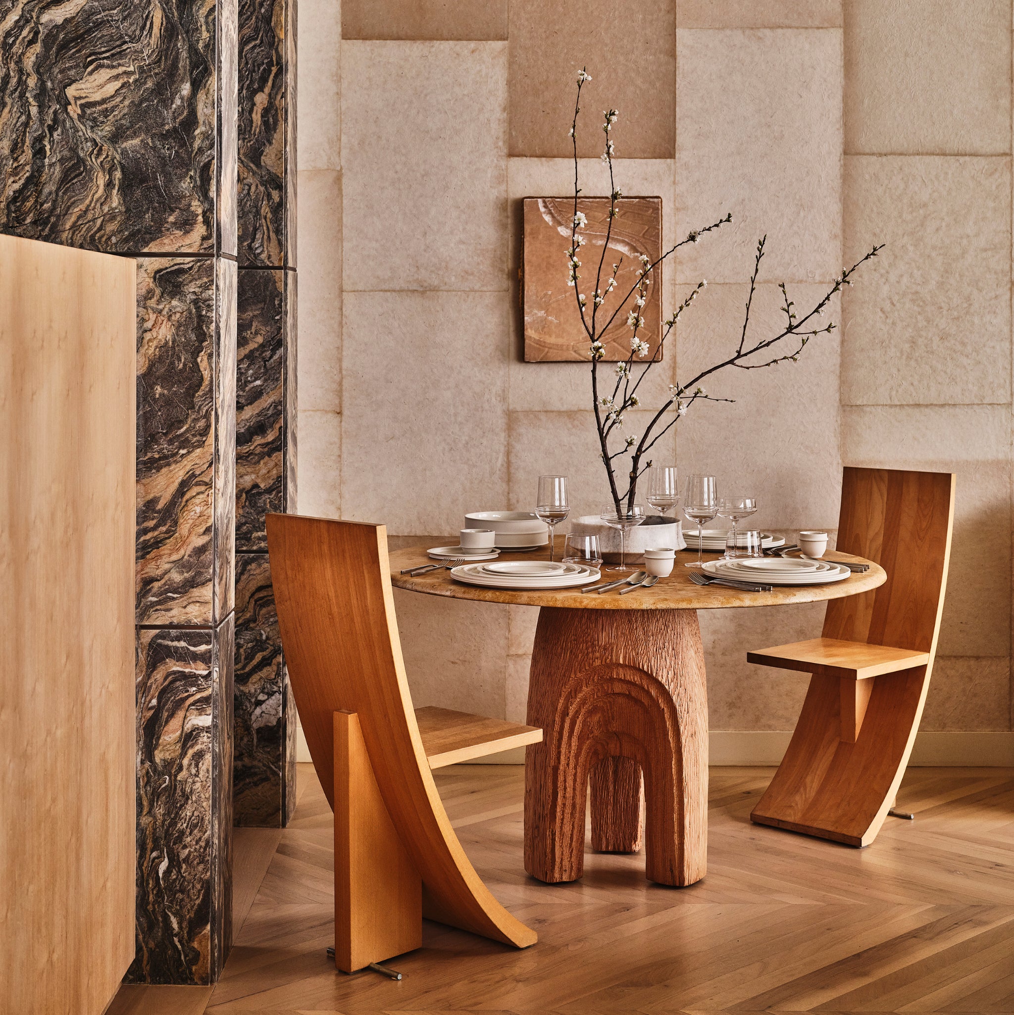 In een moderne eethoek met een ronde houten tafel en sculpturale stoelen staan Serax Dune Laag Bord Clay D17 borden op tafel en een hoge vaas met bloeiende takken, wat een door Kelly Wearstler geïnspireerde elegantie toevoegt aan de neutrale omgeving.