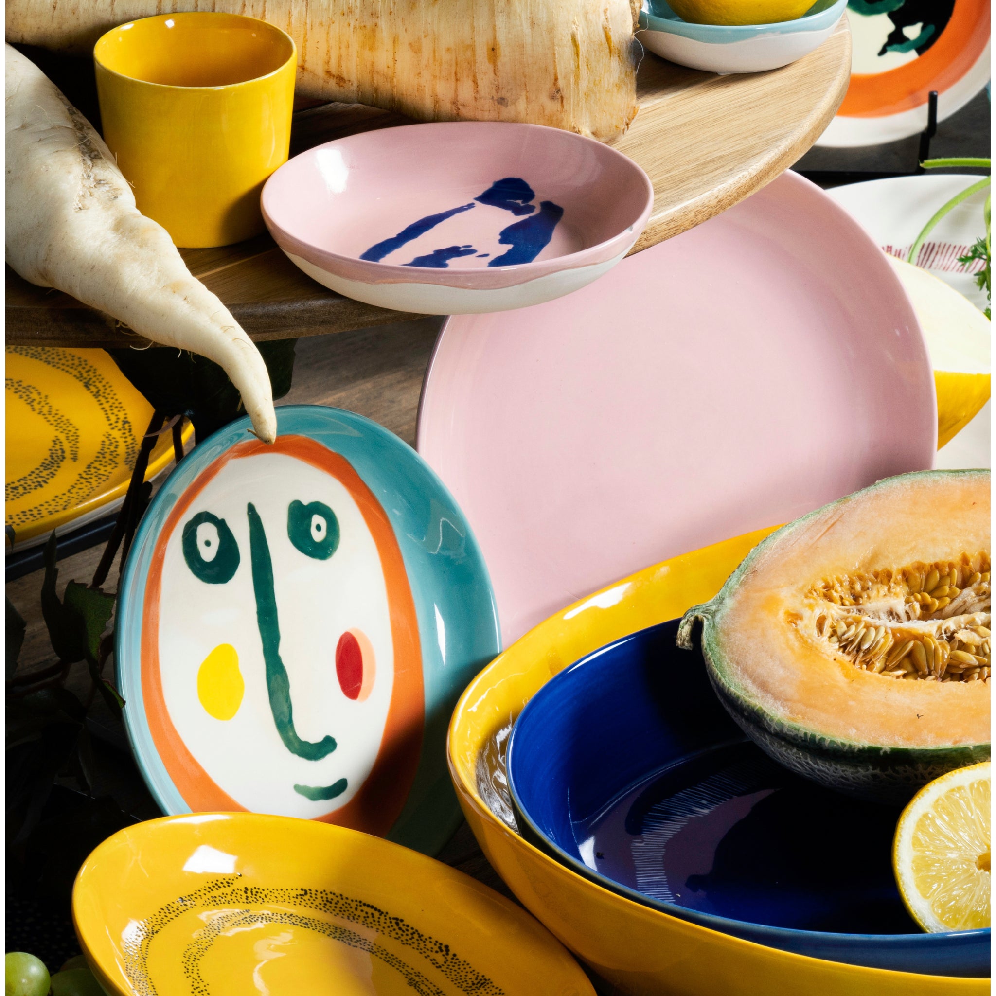 Kleurrijke keramische schalen, waaronder borden en kommen, zijn gevuld met groenten zoals meloen, citroen en pastinaak. Op één bord – het Serax Feast Ottolenghi Laag Bord Face N°1 D16 van Serax – staat een speels, abstract gezichtmotief in felle kleuren.