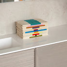 Een netjes opgevouwen Suite 702 Gastendoekjes Set C 30/50 (3 st.) van Suite 702, gemaakt van biologisch katoen met op Karel Martens geïnspireerde kleurrijke strepen, ligt op een wit aanrecht naast een wastafel met lichtgrijze stenen en houten kastjes.