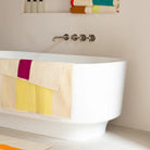 Een modern wit bad heeft een Suite 702 Badhanddoek Geel Fuchsia 70/140 gedrapeerd over de zijkant. Op een plank erboven liggen opgerolde handdoeken van biologisch katoen, met flessen netjes in de buurt.