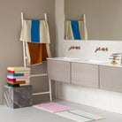 Een moderne badkamer met een dubbele wastafel, wandspiegel en een opgevouwen Suite 702 Badhanddoek Aqua Toffee 70/140 op een marmeren blok, aangevuld met een witte ladder met gedrapeerde handdoeken en een gestreepte badmat op de vloer.