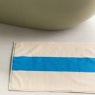 De Suite 702 Badmat Aqua Donkergroen 50/80, een crèmekleurige badmat van biologisch katoen met een opvallende blauwe streep geïnspireerd door Karel Martens, ligt naast een glad, licht bad.