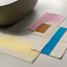 Drie badmatten van Suite 702 Badmat Aqua Donkergroen 50/80, gemaakt van biologisch katoen, liggen op een lichte vloer naast een modern bad. Hun gedurfde strepen en kleuren roepen een speelse Karel Martens vibe op.