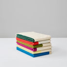 Een set van drie gevouwen Suite 702 Gastendoekjes Set D 30/50, gemaakt van biologisch katoen met kleurrijke randen, van Suite 702, weergegeven op een wit oppervlak tegen een effen grijze achtergrond. Geïnspireerd door de speelse stijl van Karel Martens.