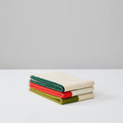 Een stapeltje van drie Suite 702 Gastendoekjes (Set D 30/50) gemaakt van biologisch katoen, met een beige hart en groene, rode en donkergroene randen, rustend op een wit oppervlak en effen grijze achtergrond.