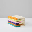 Een netjes gestapelde set van drie Suite 702 Gastendoekjes (Set C 30/50) gemaakt van biologisch katoen, elk met een verschillend gekleurde rand - oranje, geel en groen - afgebeeld op een wit oppervlak tegen een effen grijze achtergrond.