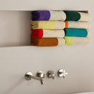 Zes netjes gevouwen Suite 702 Handdoek Donkergroen Oranje 50/100 handdoeken, met kleurrijke strepen, liggen op een verzonken lichtbeige plank boven een badkamerwastafel met een zilveren kraan en twee handgrepen.