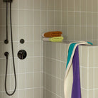 Een moderne douche met lichtgrijze vierkante tegels, zwarte douchekop en handgreep, en een halve muur bedekt met gevouwen biokatoenen handdoeken; een Suite 702 Handdoek Lime Toffee 50/100 van Suite 702 hangt over de muur.