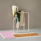 Een modern, sculpturaal handdoekenrek toont verschillende kleurrijke handdoeken, waaronder de Suite 702 Handdoek Donkergroen Oranje 50/100. Twee rechthoekige badmatten - een roze en een bruine met witte randen - liggen op een lichtgrijze vloer tegen een beige muur.