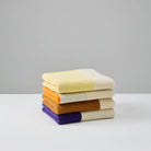 Een keurig gestapelde stapel van vier Suite 702 Handdoeken Rood Geel 50/100, met op Karel Martens geïnspireerde gele, oranje, bruine en paarse strepen, ligt op een wit oppervlak met een effen lichtgrijze achtergrond.