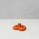 De Wavy Kandelaar Rood van Wavy Design Studio is een kleine, geribbelde oranje vaas van keramiek met drie ronde bodems en een centrale opening, die op een licht oppervlak tegen een grijze betonnen muur staat.