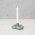 Een witte conische kaars staat in de Wavy Kandelaar Muntgroen van Wavy Design Studio, een moderne lichtgroene kandelaar met drie onderling verbonden ringen, op een licht oppervlak tegen een betonnen muur.