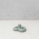 De Wavy Kandelaar Muntgroen van Wavy Design Studio, een minimalistische golvende kandelaar, staat op een lichtgekleurd oppervlak naast een lichtgroene ronde vaas met geribbelde textuur, tegen een betonnen muur.