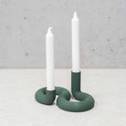 Twee witte kaarsen in de Wavy Dubbele Kandelaar Highland Groen van Wavy Design Studio voegen een vleugje duurzaam interieur toe aan een licht oppervlak en steken af tegen een grijze, gespikkelde achtergrond.