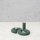 Twee geribbelde, gebogen groene kandelaars - de Wavy Dubbele Kandelaar Highland Groen van Wavy Design Studio - staan op een licht gestructureerd oppervlak tegen een betonnen muur. De ene staat hoog en rechtop, de andere is korter en meer opgerold, perfect voor duurzaam interieur.