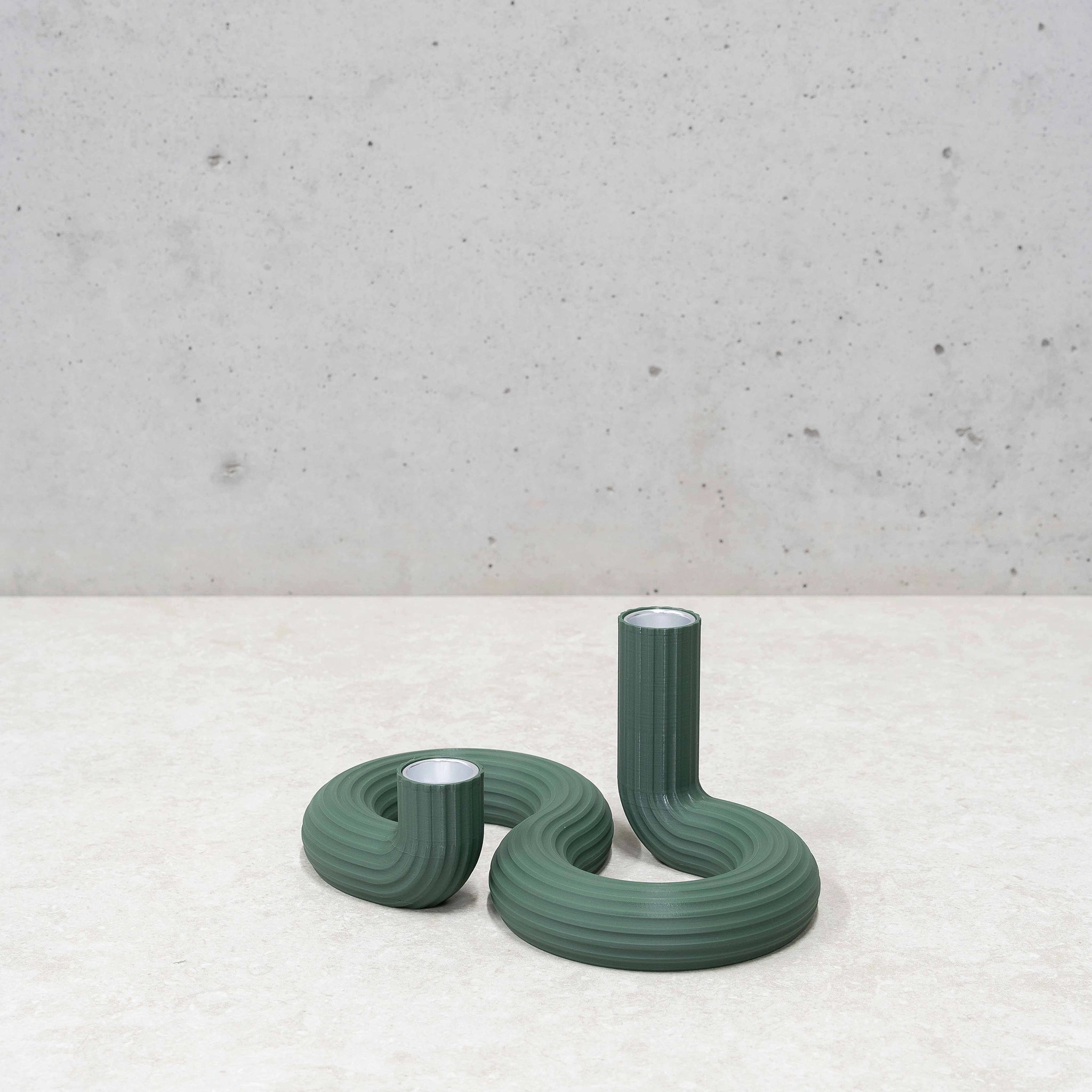 Twee geribbelde, gebogen groene kandelaars - de Wavy Dubbele Kandelaar Highland Groen van Wavy Design Studio - staan op een licht gestructureerd oppervlak tegen een betonnen muur. De ene staat hoog en rechtop, de andere is korter en meer opgerold, perfect voor duurzaam interieur.